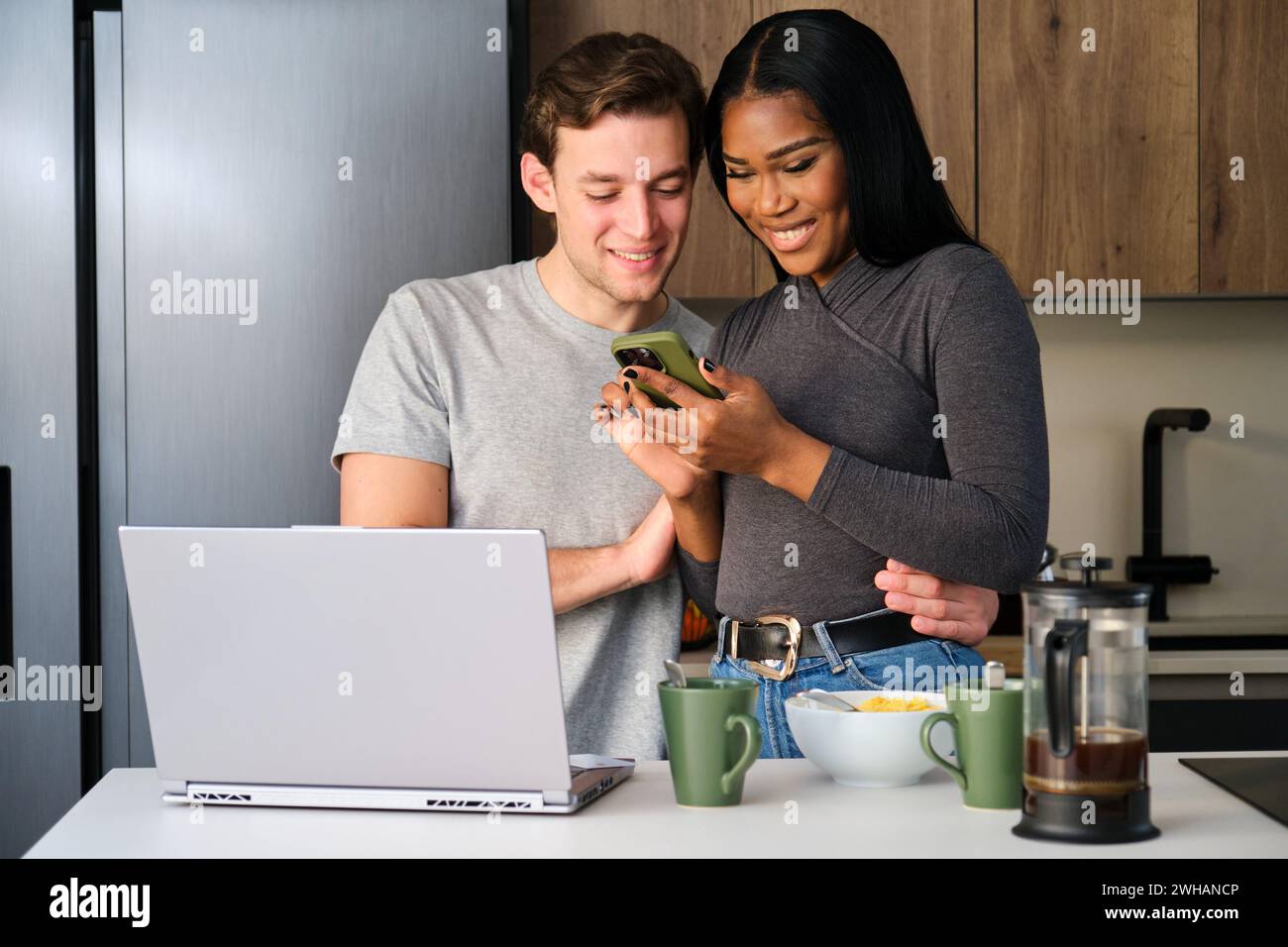Multiethnic couple using the phone while having breakfast at kitchen. Stock Photo