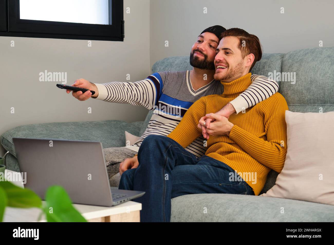 Homosexual happy couple watching TV, turning on with remote control. Stock Photo