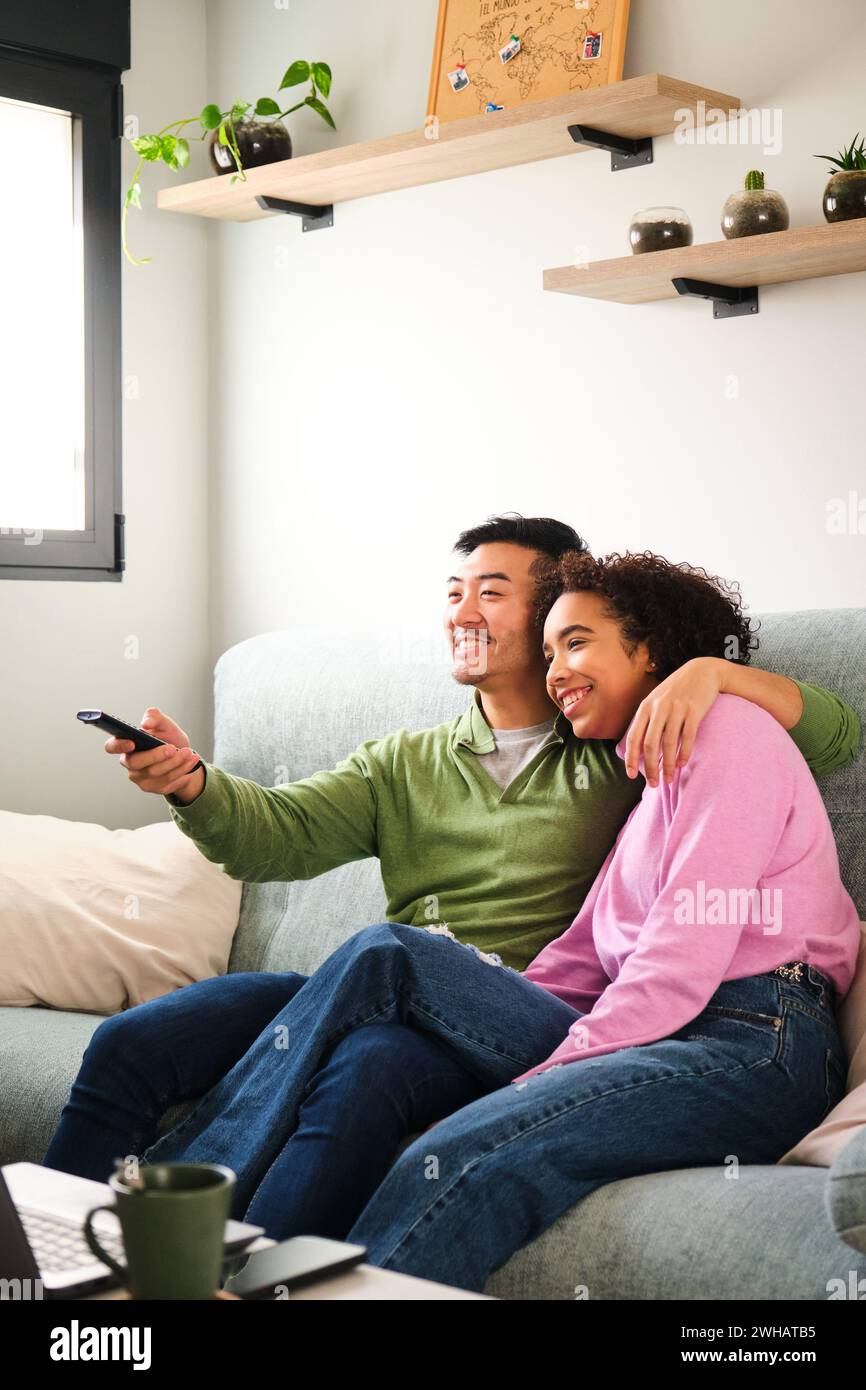 Multiracial couple smile and watch TV, movie or film together on sofa. Stock Photo