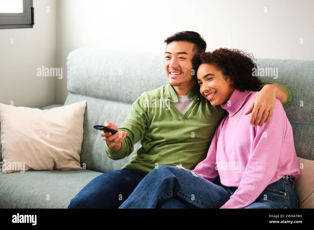 Multiethnic couple smile and watch TV, movie or film together on sofa. Stock Photo