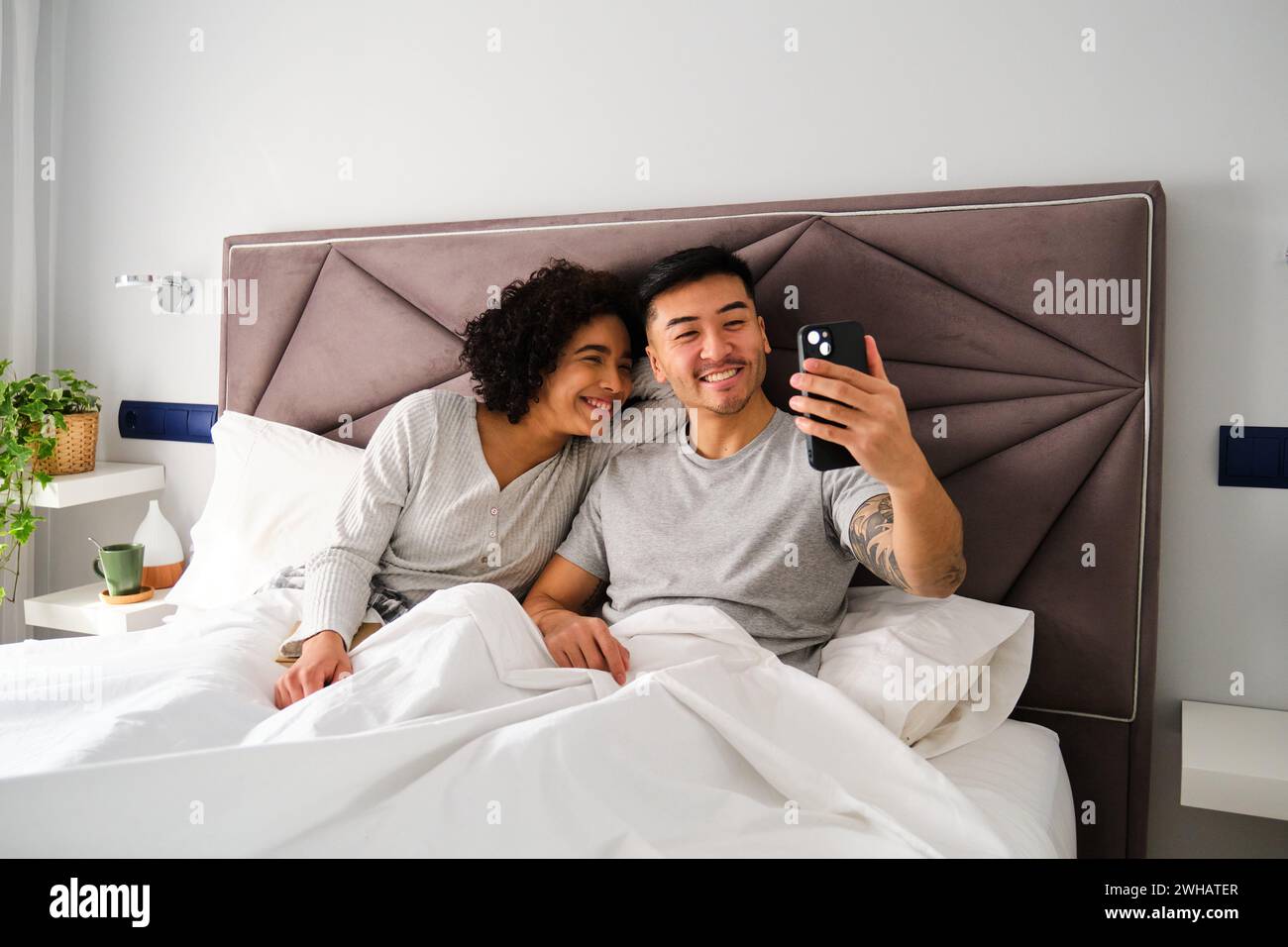 Multiethnic couple smiling and taking a selfie on bed. Stock Photo