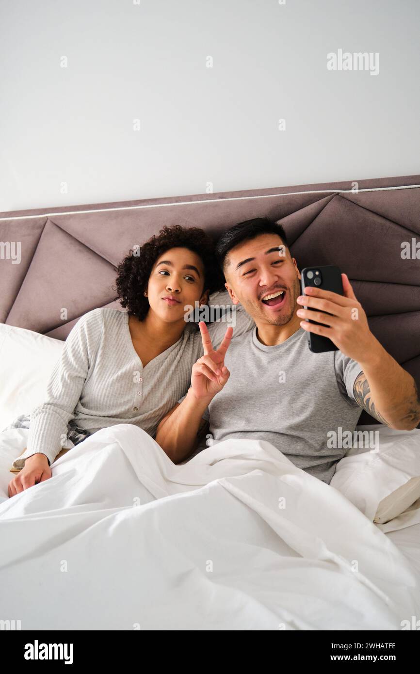 Diverse couple smiling and taking a selfie on bed. Stock Photo