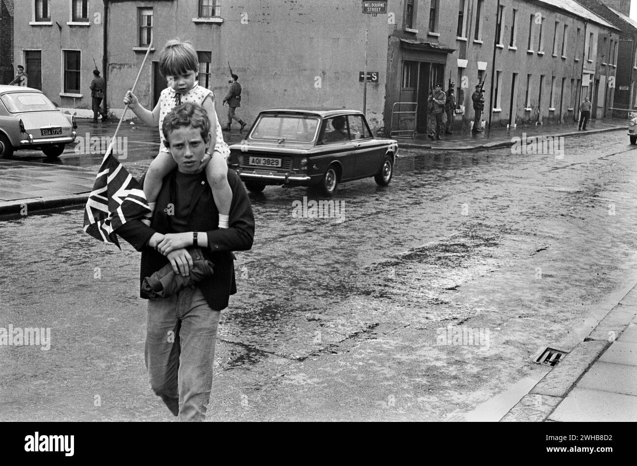 1970s Belfast. a young boy and his sister who is carrying a Union Jack flag return from an Orange Day parade. Walking through depressed back streets, while the British army patrol this inner city area. Northern Ireland. 1970 HOMER SYKES Stock Photo