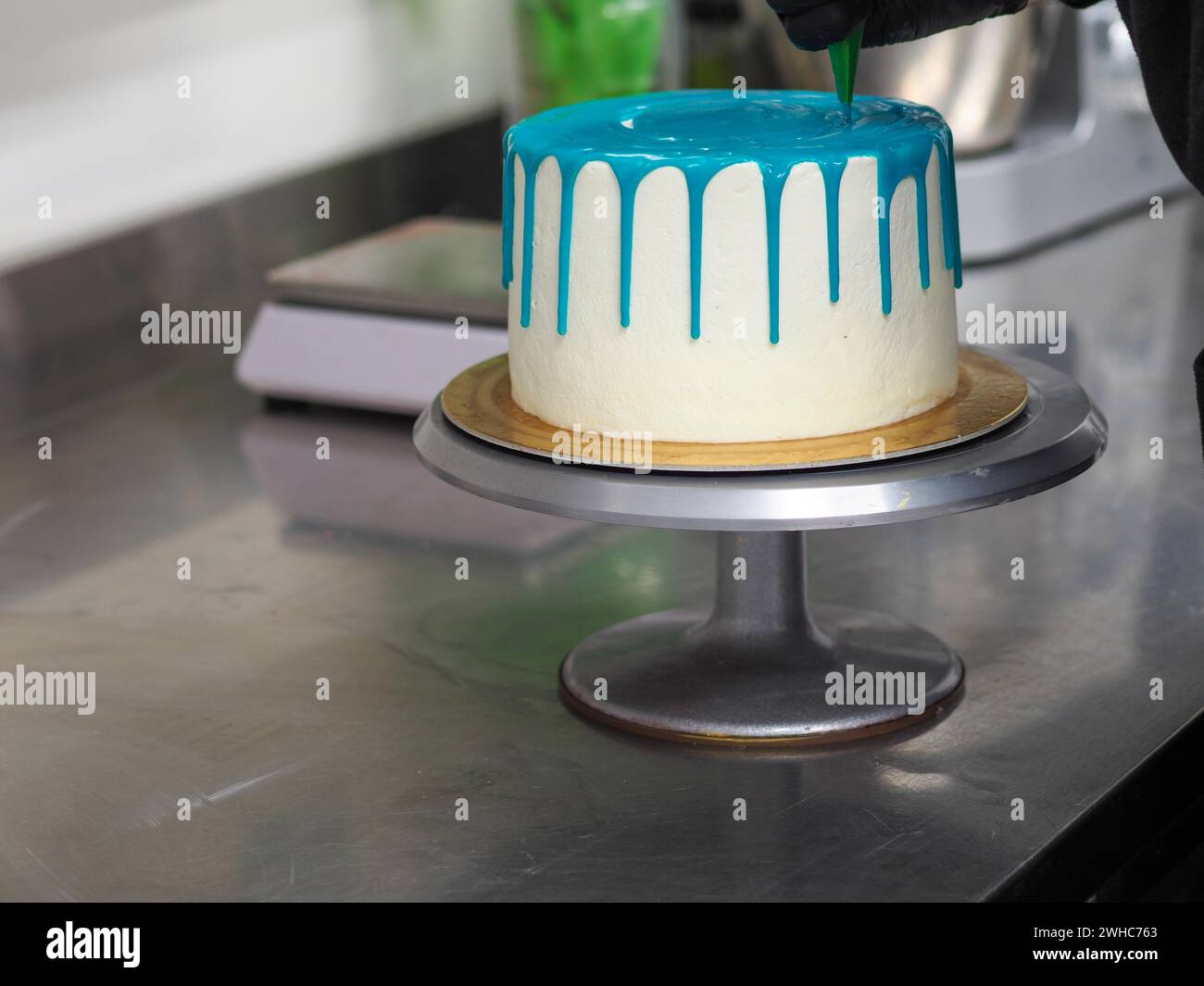 A frosted cake with white frosting being decorated with blue icing on a kitchen counter by pastry chef wearing black latex gloves Stock Photo
