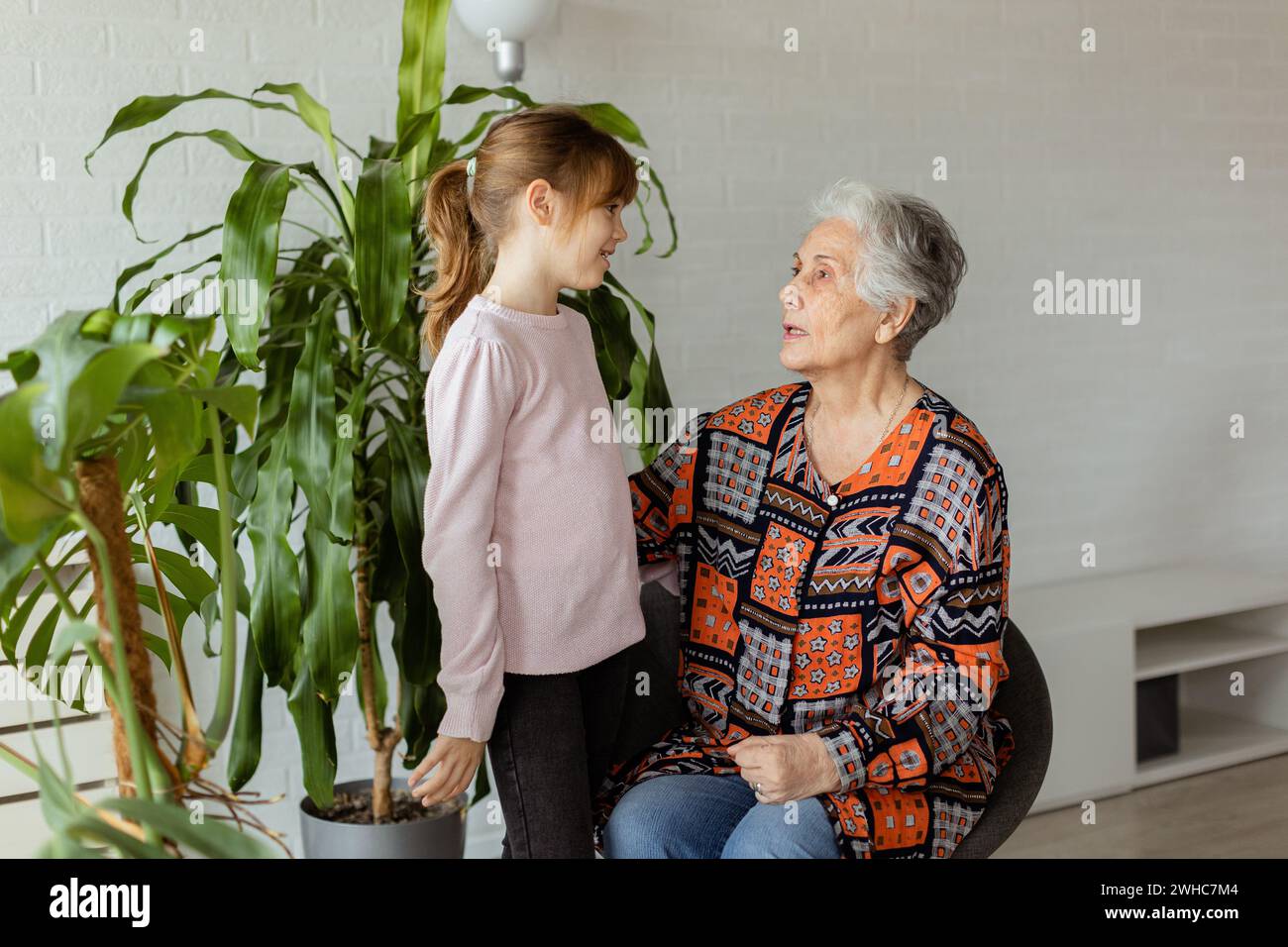 In a softly-lit room, a young girl stands beside a potted plant, sharing a moment of connection and conversation with her elderly grandmother, the joy Stock Photo