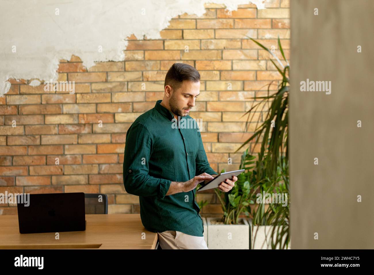 Cheerful man stands confidently holding a digital tablet in a contemporary office space with an exposed brick wall, symbolizing a blend of modern tech Stock Photo