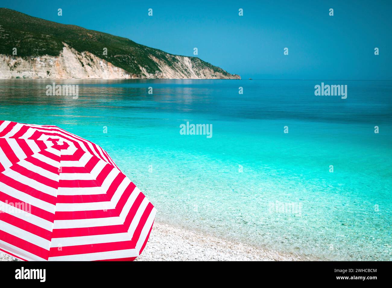Striped red sun beach umbrella on a pebble beach with turquoise blue sea, white rocks and sky in background. Stock Photo