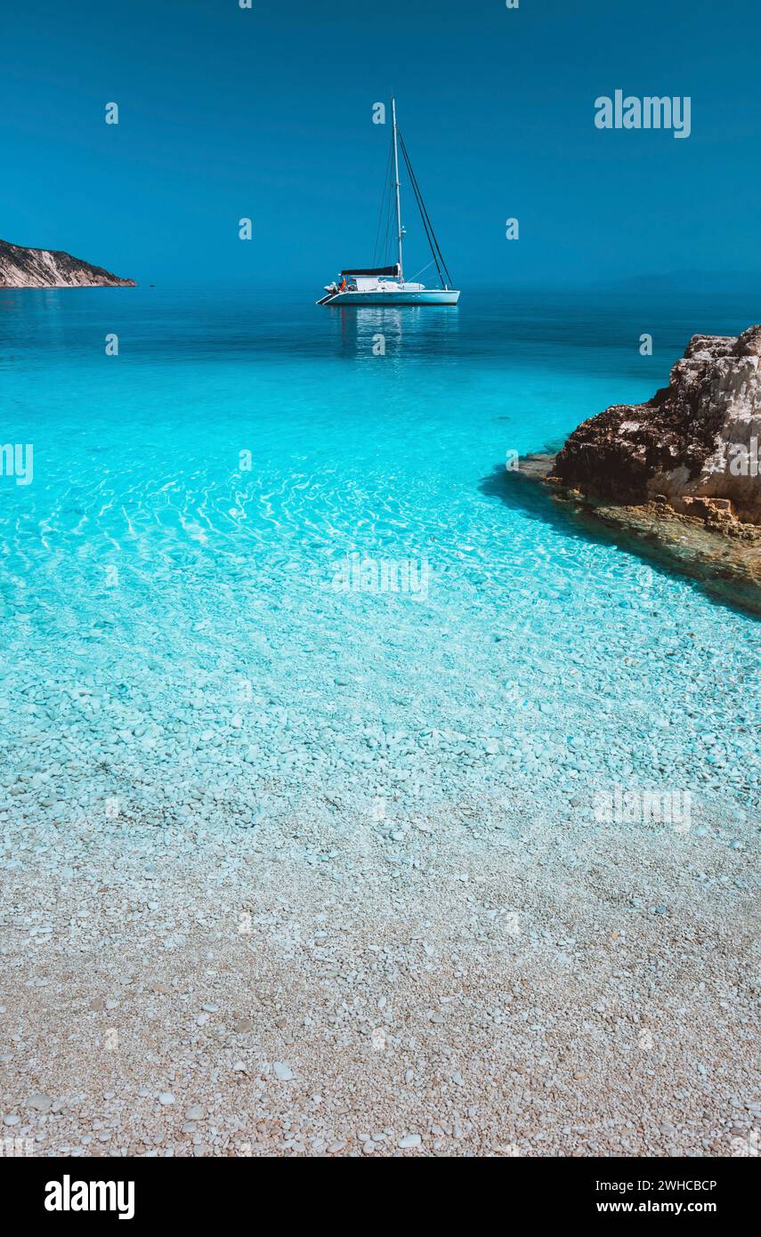 Lonely white sailing catamaran boat drift on calm sea surface. Pure azure clean blue lagoon with shallow water and pebble beach. Some brown rock stones in foreground. Stock Photo