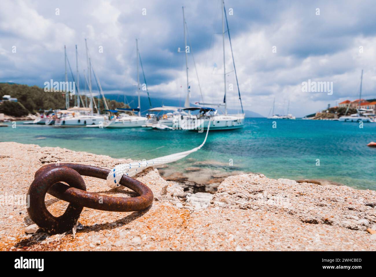 Mooring rope tied to rusty ring for rigging yachts in background. Wonderful view of port Fiskardo. Picturesque seascape of Ionian Sea. Outdoor scene of Kefalonia island, Greece, Europe. Stock Photo