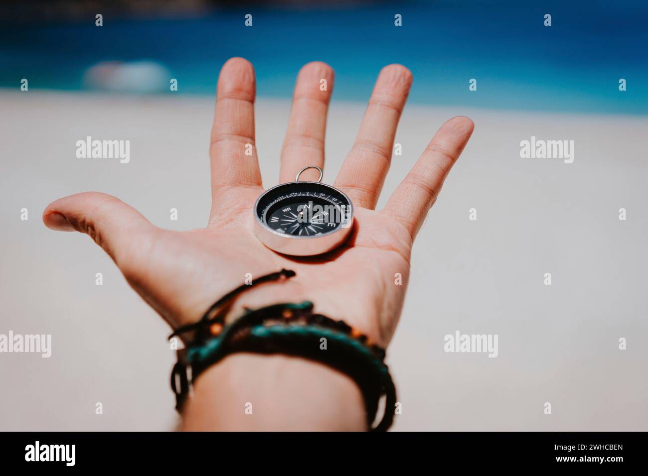 Open palm with stretched fingers holding black metal compass against white sandy beach. Find your way or goal concept. Point of view pov. Stock Photo