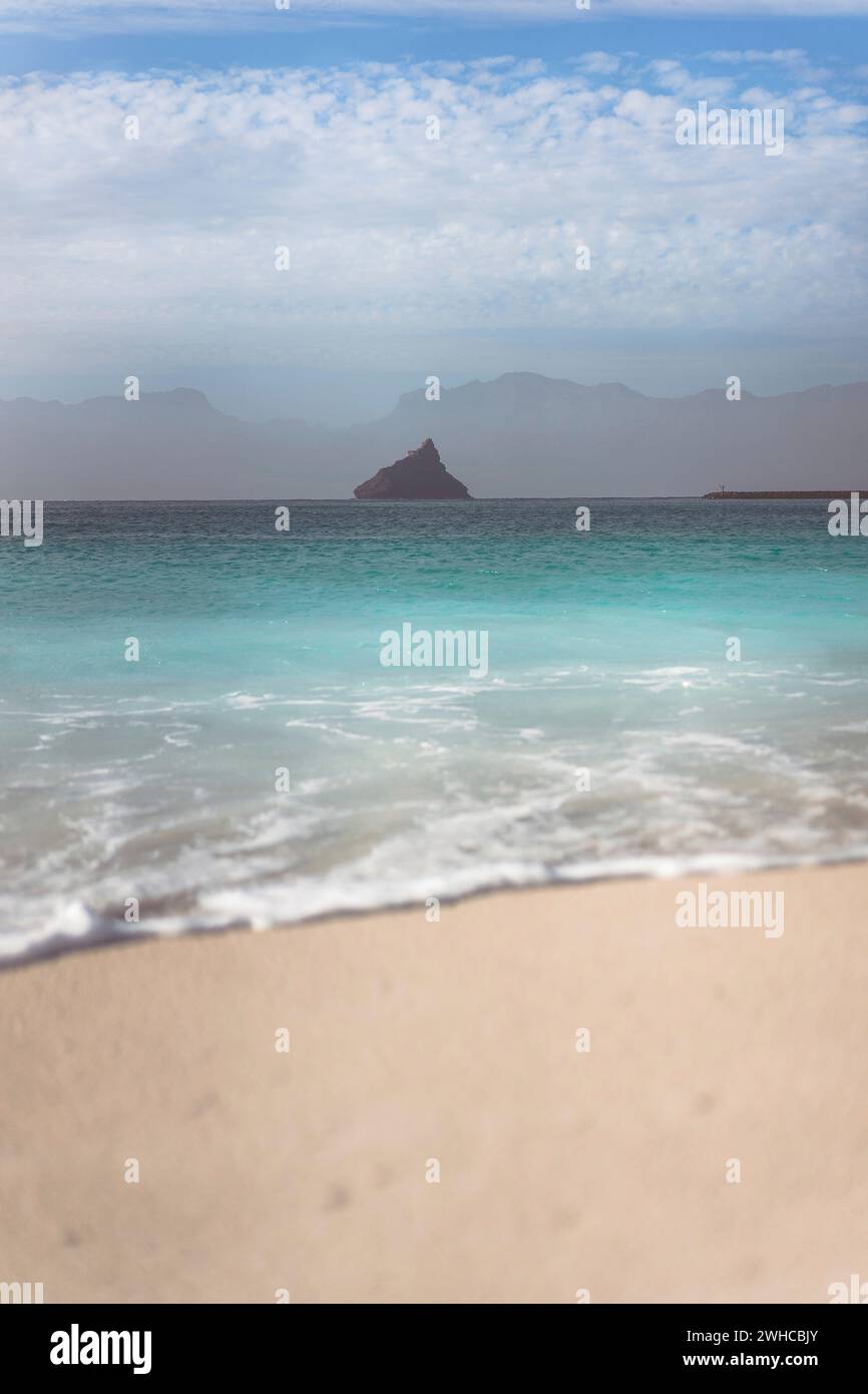 Black rock on ocean horizon. Defocused ocean wave rolling to sandy beach with clear transparent azure water. White clouds in sky. Stock Photo