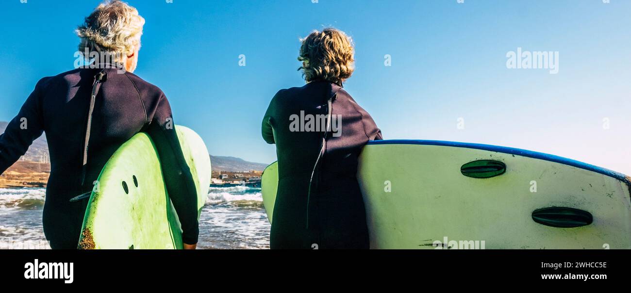 couple of seniors at the beach with black wetsuits holding a surftable ready to go surfing a the beach - active mature and retired people doing happy activity together in their vacations or freetime Stock Photo