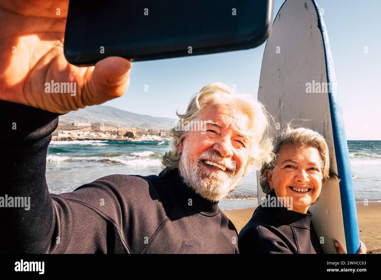 couple of pensioners seniors taking a selfie together at the beach with their wetsuits and surfboards - mature people learning surfing Stock Photo