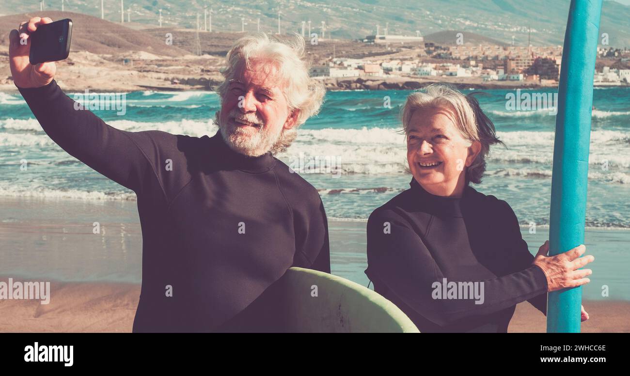 happy couple of seniors at the beach trying to go surf and having fun together - mature woman and man married taking a selfie with the wetsuits and surftables with sea or ocean at the background Stock Photo