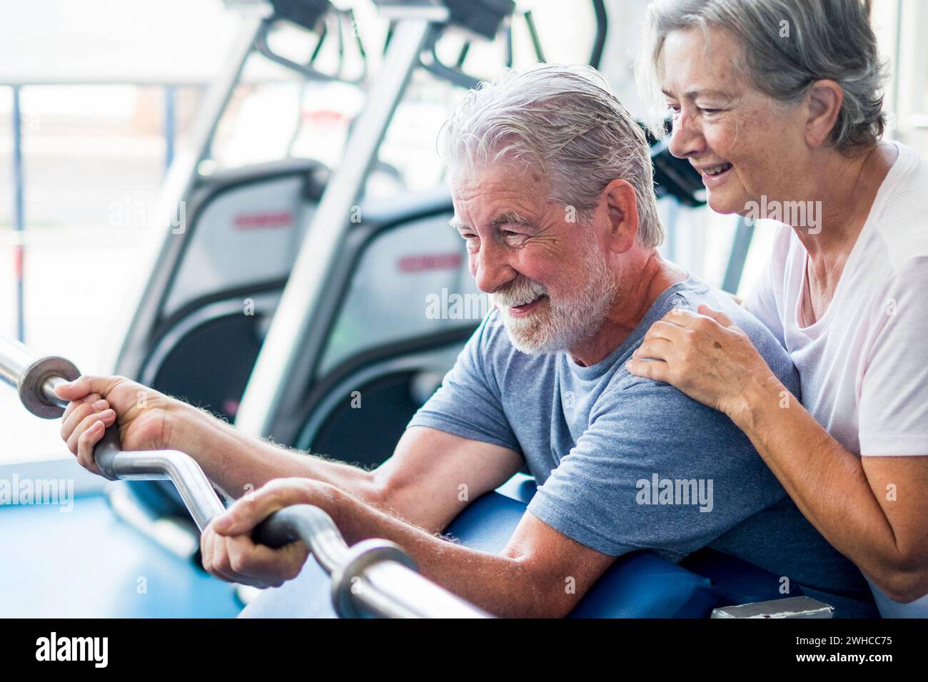 beautiful couple of two seniors at the gym doing exercises with a strong light at the background - woman hugging her husband holding a barr - active lifestyle Stock Photo