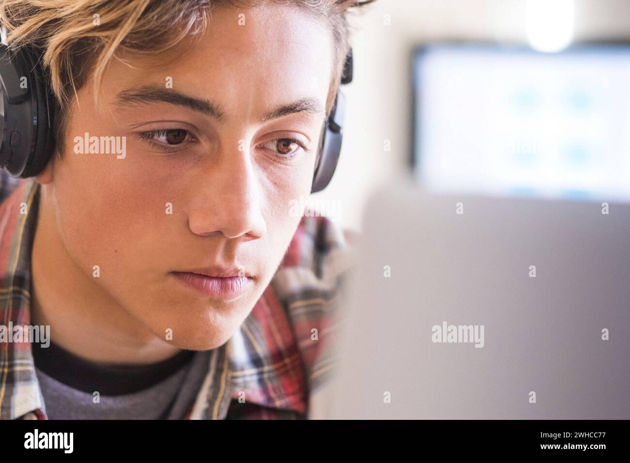 caucasian teenager indoor doing homework on the table at home - blonde guy writing and reading in his laptop or computer to get greats scores - portrait of boy with headphones Stock Photo