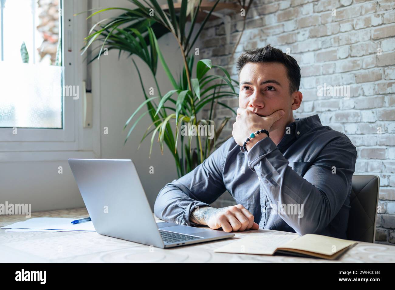 Thoughtful businessman touching chin, pondering ideas or strategy, sitting at wooden work desk with laptop, freelancer working on online project, student preparing for exam at home Stock Photo