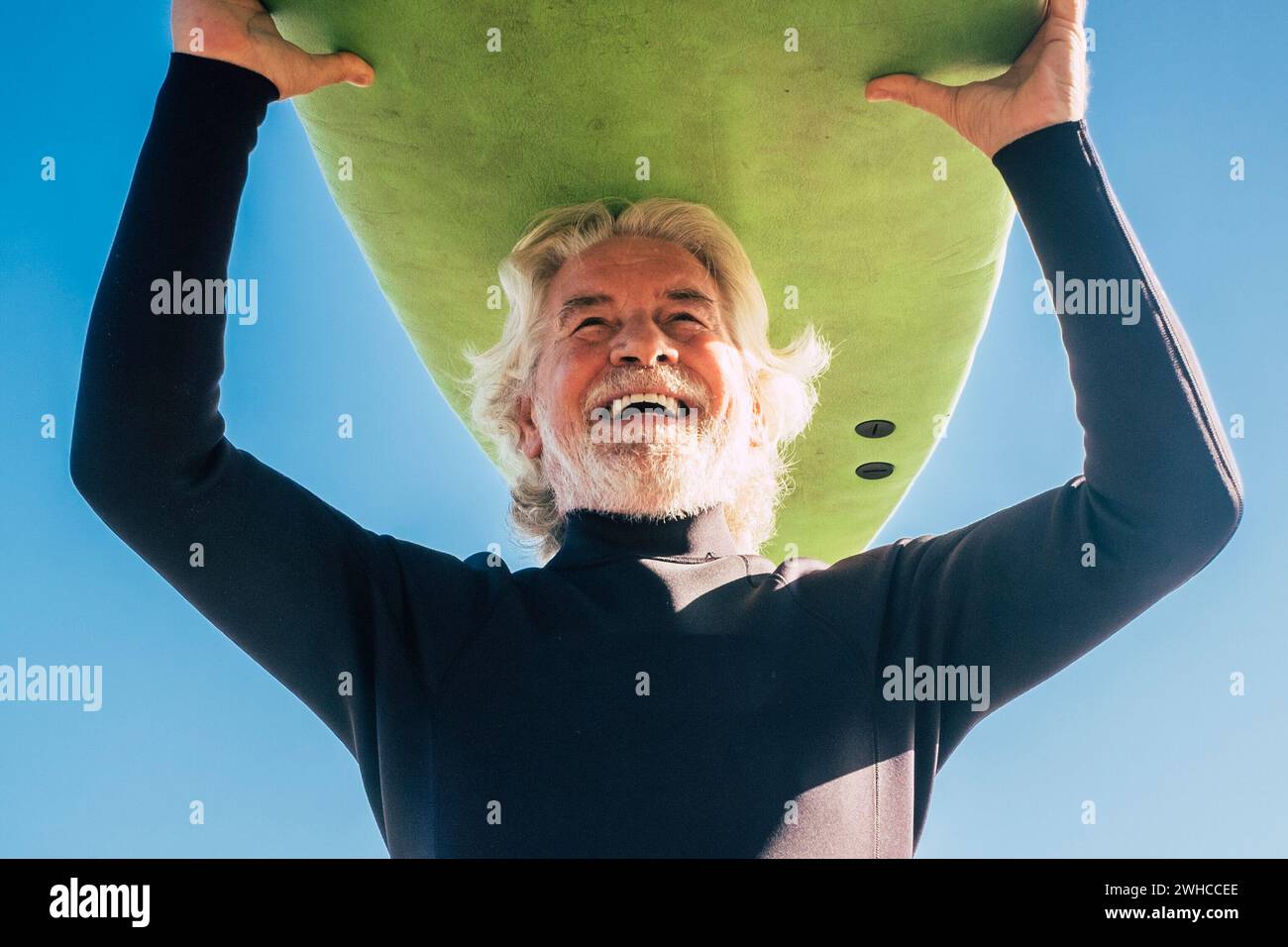 happy senior with surftable on his head is smiling and laughing - old and mature man having fun surfing with a black wetsuits - active retired adult doing activity alone Stock Photo