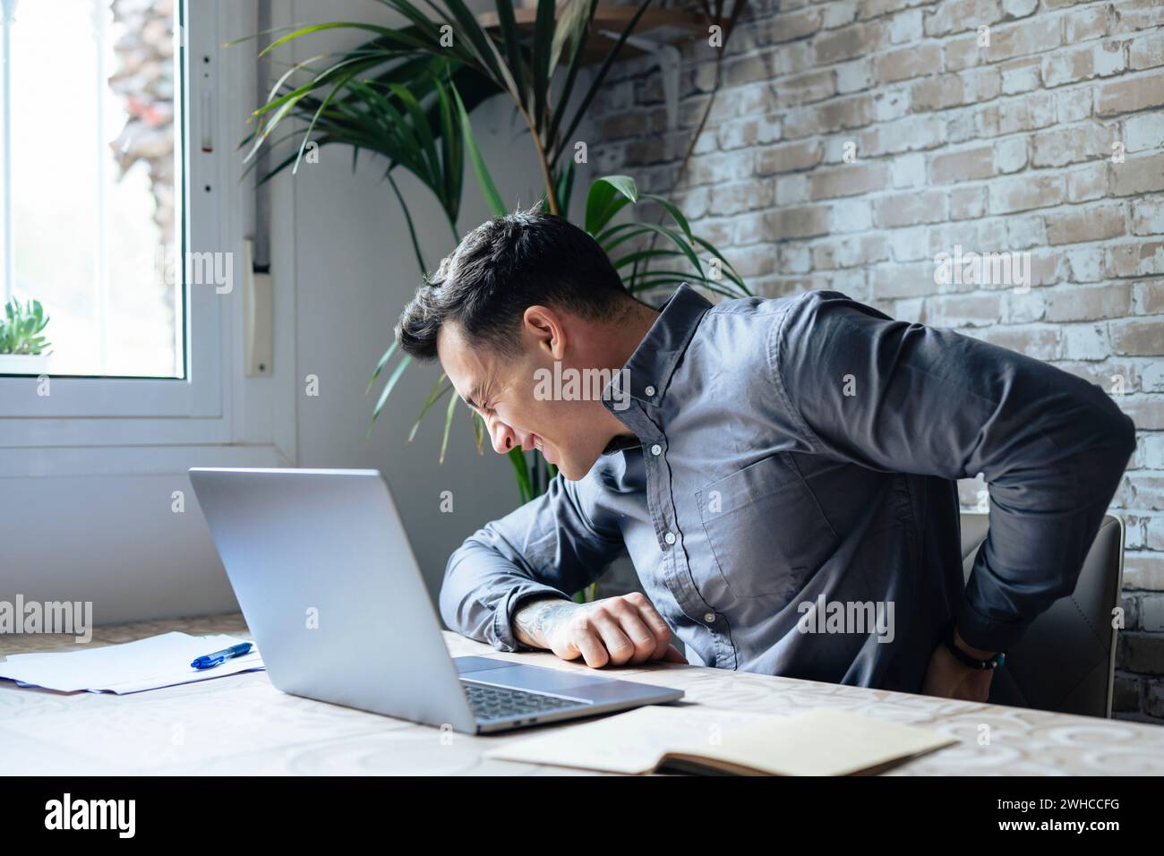 Unhappy Caucasian man sit at desk in home office work on laptop suffer from lower back muscle strain or spasm. Unwell unhealthy male have backache from incorrect posture. Sedentary life concept. Stock Photo