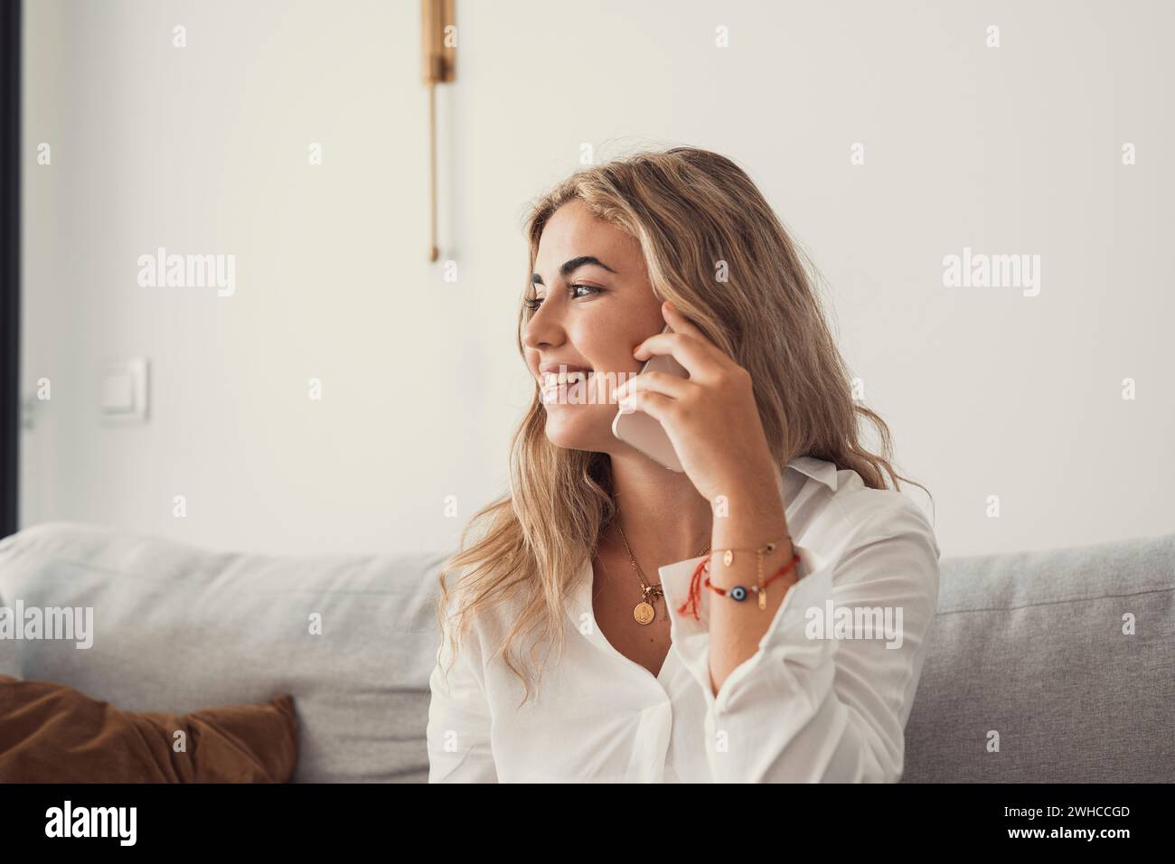 Happy cheerful young woman talking on the phone at home, smiling teen girl making answering call by cellphone sitting on couch, beautiful lady having pleasant funny conversation speaking by mobile Stock Photo