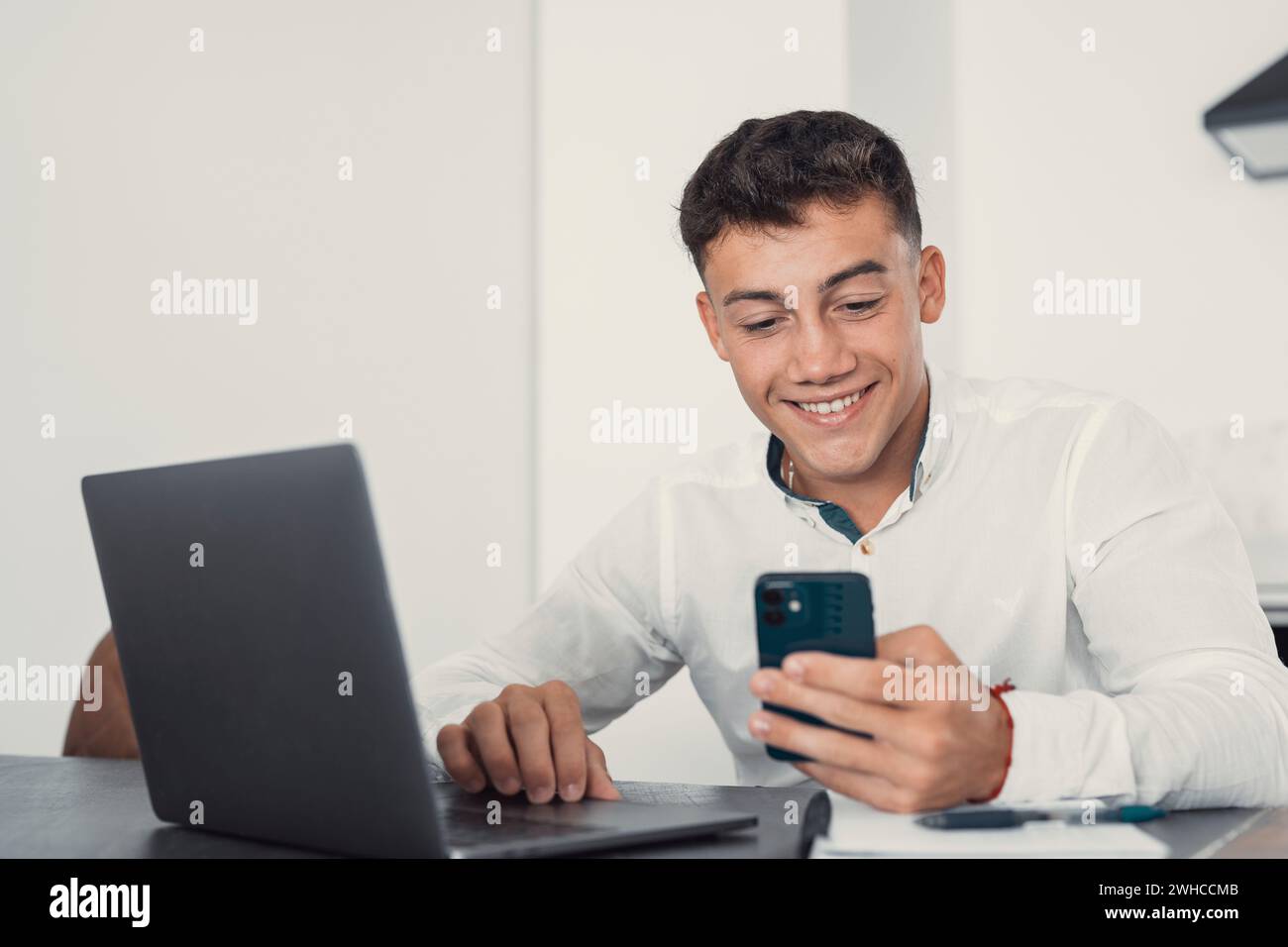 Confident man sitting at desk taking break in work with electronic documents on laptop to make answer telephone call. Smiling young guy freelancer synchronize data between home computer and smartphone Stock Photo