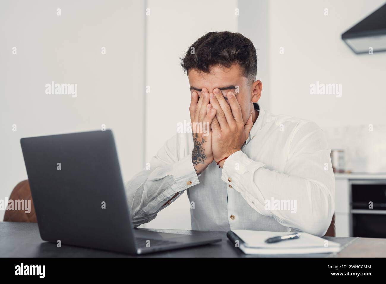 Tired young man feel pain eyestrain holding glasses rubbing dry irritated eyes fatigued from computer work, stressed man suffer from headache bad vision sight problem sit at home table using laptop Stock Photo