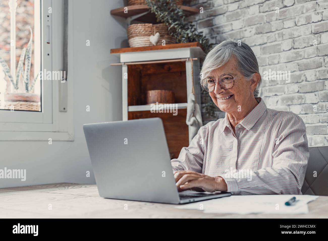 Aged remote worker. Concentrated senior female in glasses work on laptop from home office read email electronic document. Old age woman employee freelancer sit at kitchen table by pc typing report online Stock Photo