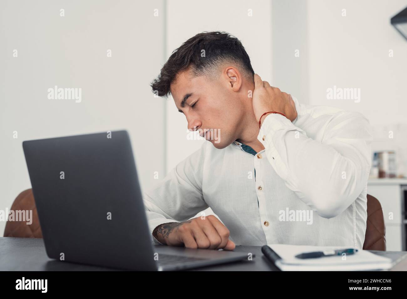 Exhausted young Caucasian male worker sit at desk massage neck suffer from strain spasm muscles. Tired unwell man overwhelmed with computer work sedentary lifestyle struggle with back pain or ache. Stock Photo
