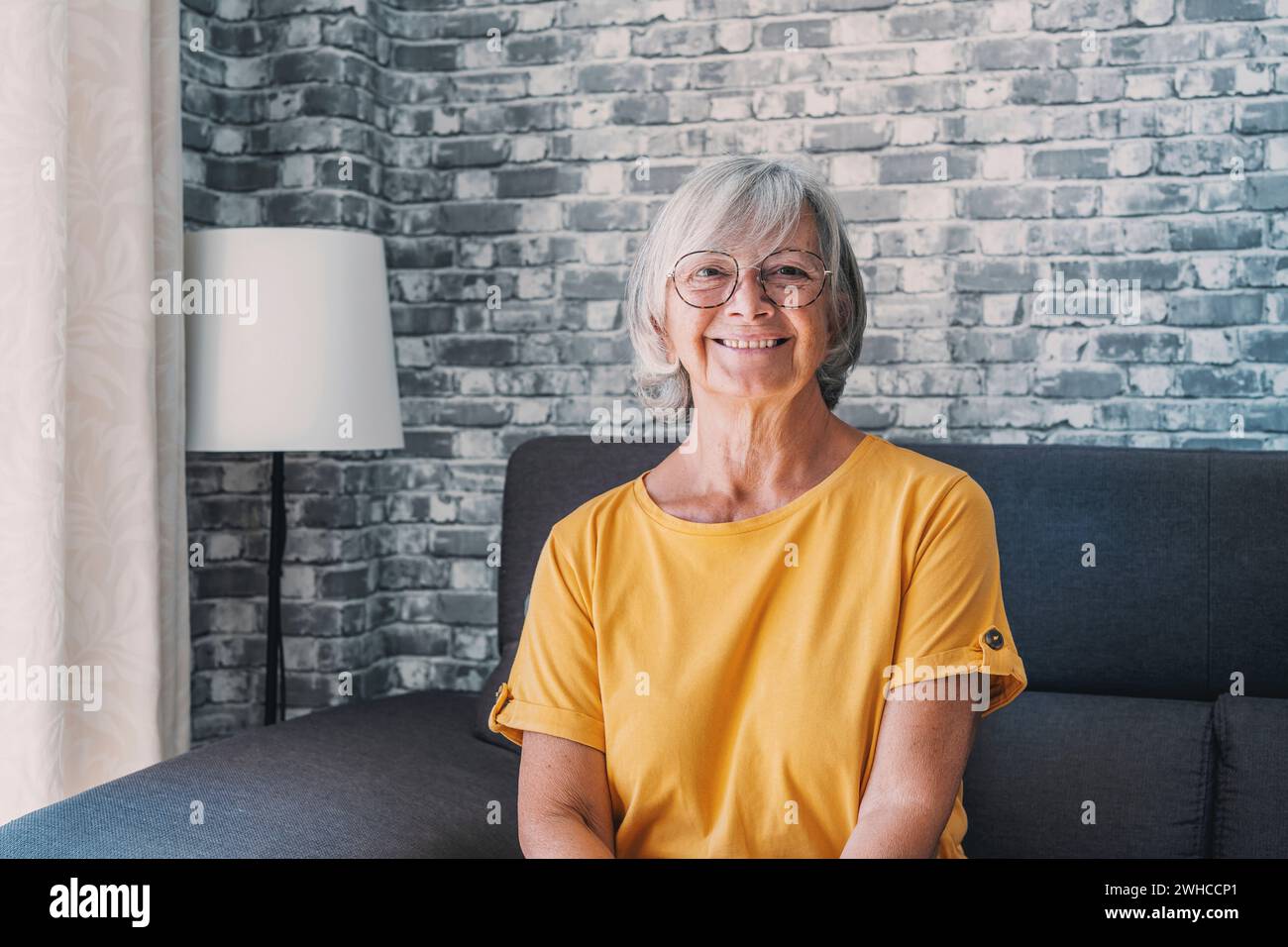 Smiling middle aged mature grey haired woman looking at camera, happy old lady in glasses posing at home indoor, positive single senior retired female sitting on sofa in living room headshot portrait Stock Photo