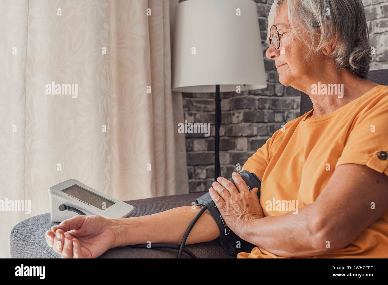 Senior elder woman measure high low blood pressure test holding using medical digital electronic meter sit on sofa at home, heart disease atherosclerosis prevention, hypotension hypertension concept Stock Photo
