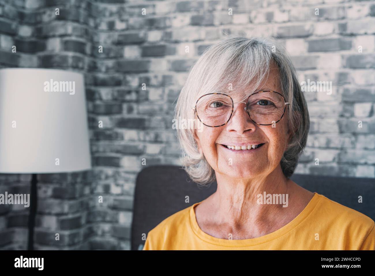 Smiling middle aged mature grey haired woman looking at camera, happy old lady in glasses posing at home indoor, positive single senior retired female sitting on sofa in living room headshot portrait Stock Photo