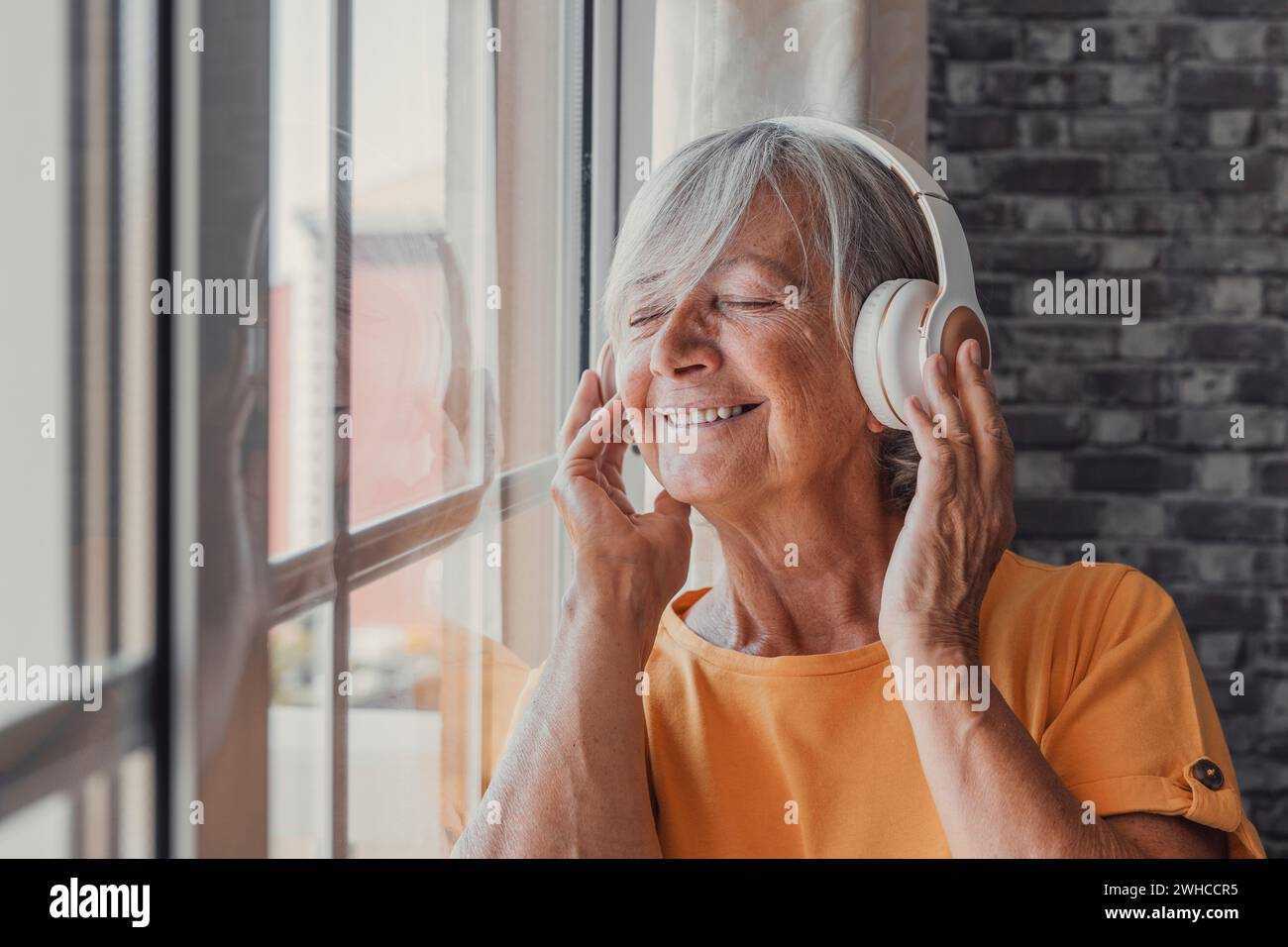Side view happy old mature retired woman listening popular music in headphones, relaxing alone looking thought window, enjoying peaceful carefree weekend time alone at home, stress free leisure pastime. Stock Photo