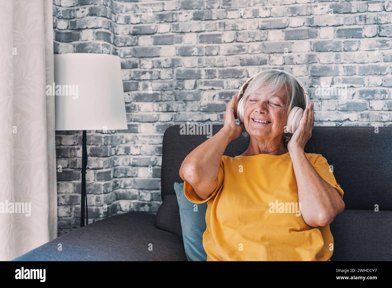 Side view happy old mature retired woman listening popular music in headphones, relaxing alone on cozy sofa, enjoying peaceful carefree weekend time alone at home, stress free leisure pastime. Stock Photo