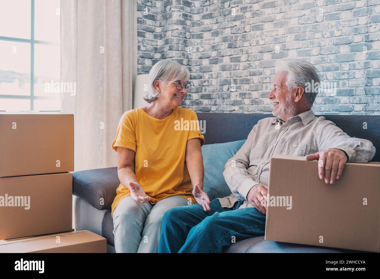 Relaxed mature married couple in love resting on couch among paper cardboard boxes, taking break, pause, hugging, talking, enjoying being in new home. Real estate concept Stock Photo