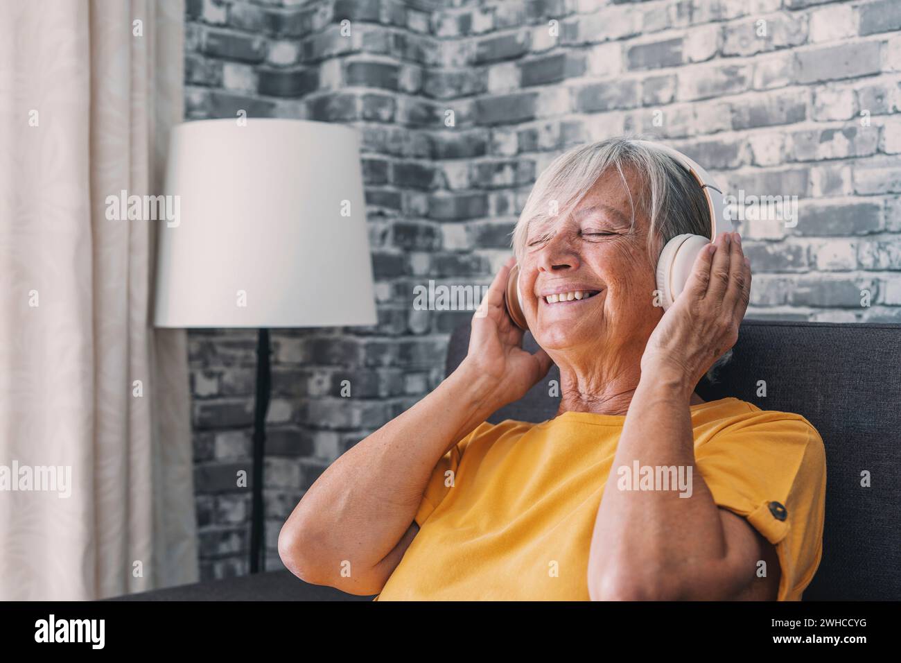 Side view happy old mature retired woman listening popular music in headphones, relaxing alone on cozy sofa, enjoying peaceful carefree weekend time alone at home, stress free leisure pastime. Stock Photo