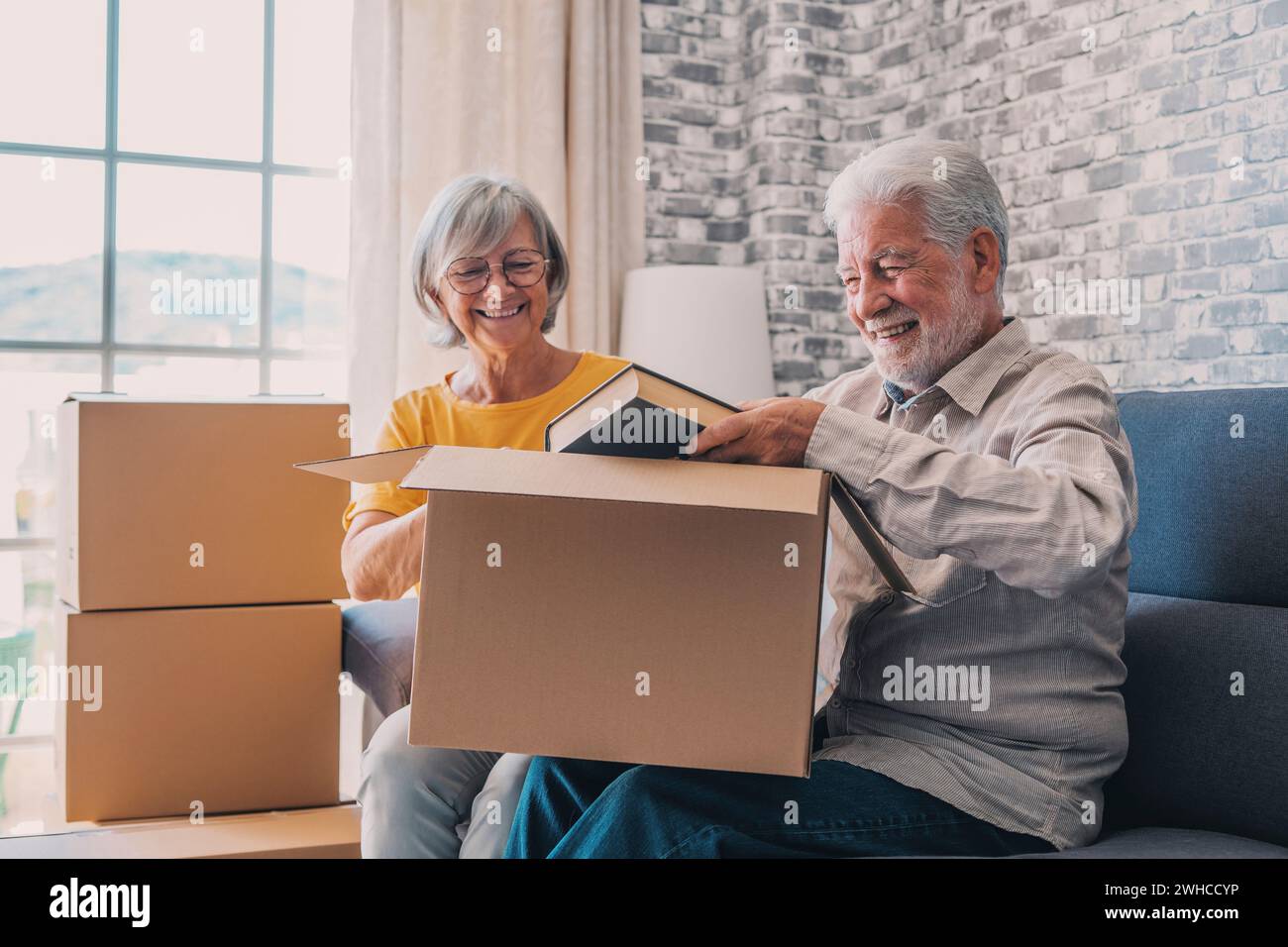 Relaxed mature married couple in love resting on couch among paper cardboard boxes, taking break, pause, hugging, talking, enjoying being in new home. Real estate concept Stock Photo
