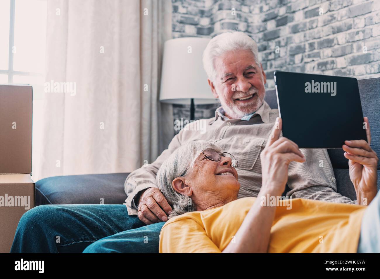 Happy retired senior family couple relaxing on sofa after moving activities, sharing tablet computer, looking at screen, laughing, making video call, using online app, Internet service Stock Photo
