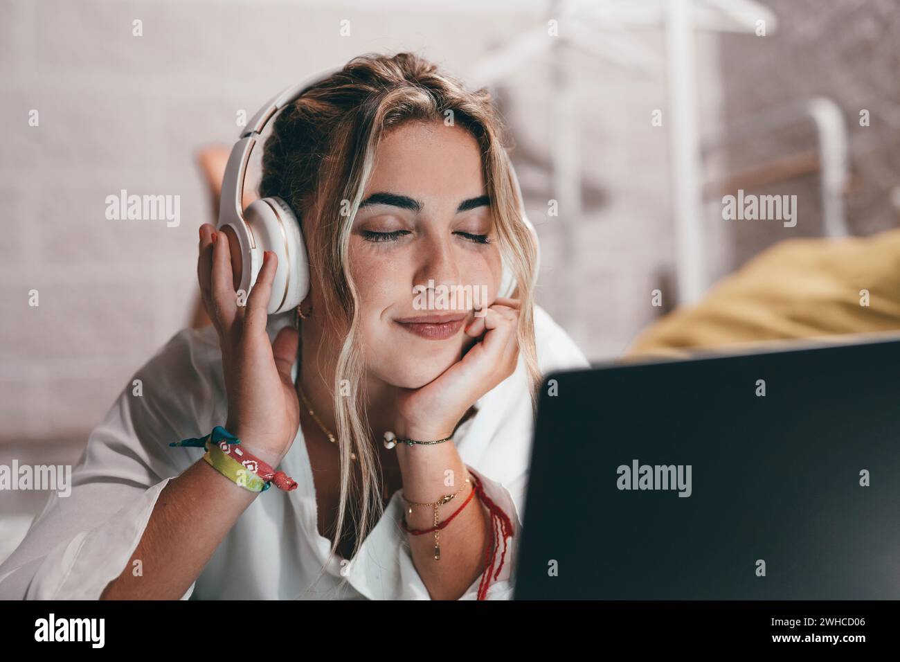 Close up of one young pretty happy woman using laptop indoor at home lying on the bed wearing headphones listening music. Millennial teenager relaxing enjoying songs with eyes closed Stock Photo