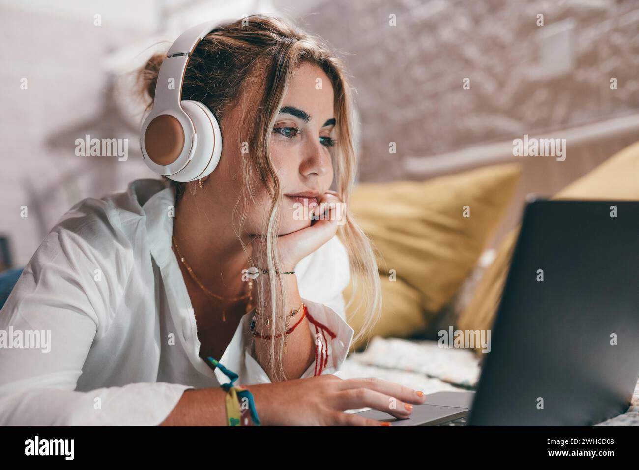 Close up of one young pretty happy woman using laptop indoor at home lying on the bed wearing headphones listening music. Millennial teenager having fun online studying. Stock Photo