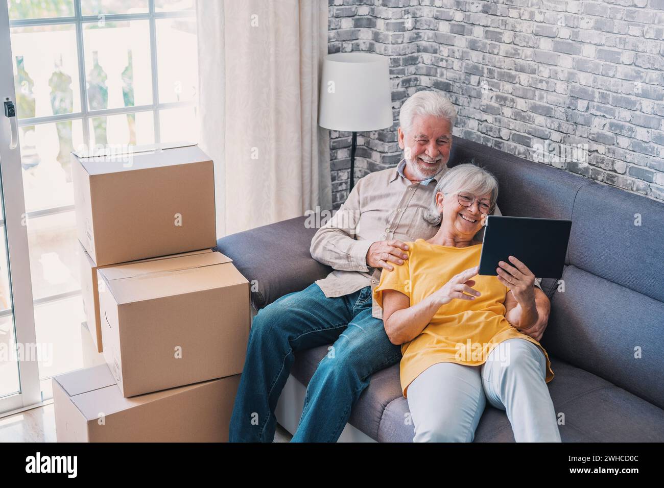 Happy retired senior family couple relaxing on sofa after moving activities, sharing tablet computer, looking at screen, laughing, making video call, using online app, Internet service Stock Photo