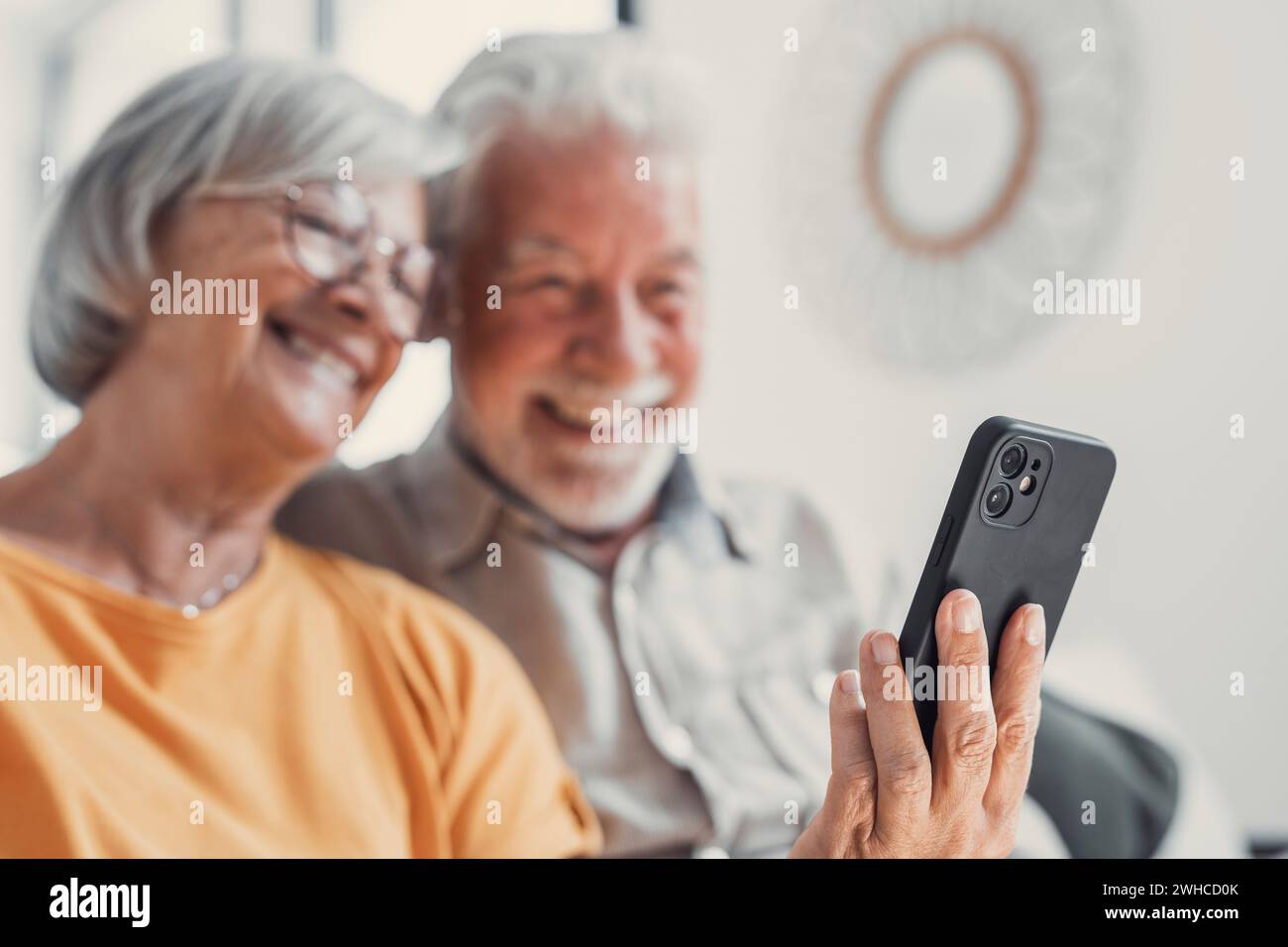 Happy mature family, wife and husband using phone together at home, smiling middle aged man and woman sitting on couch, using mobile device apps, watching video in social network, surfing internet Stock Photo