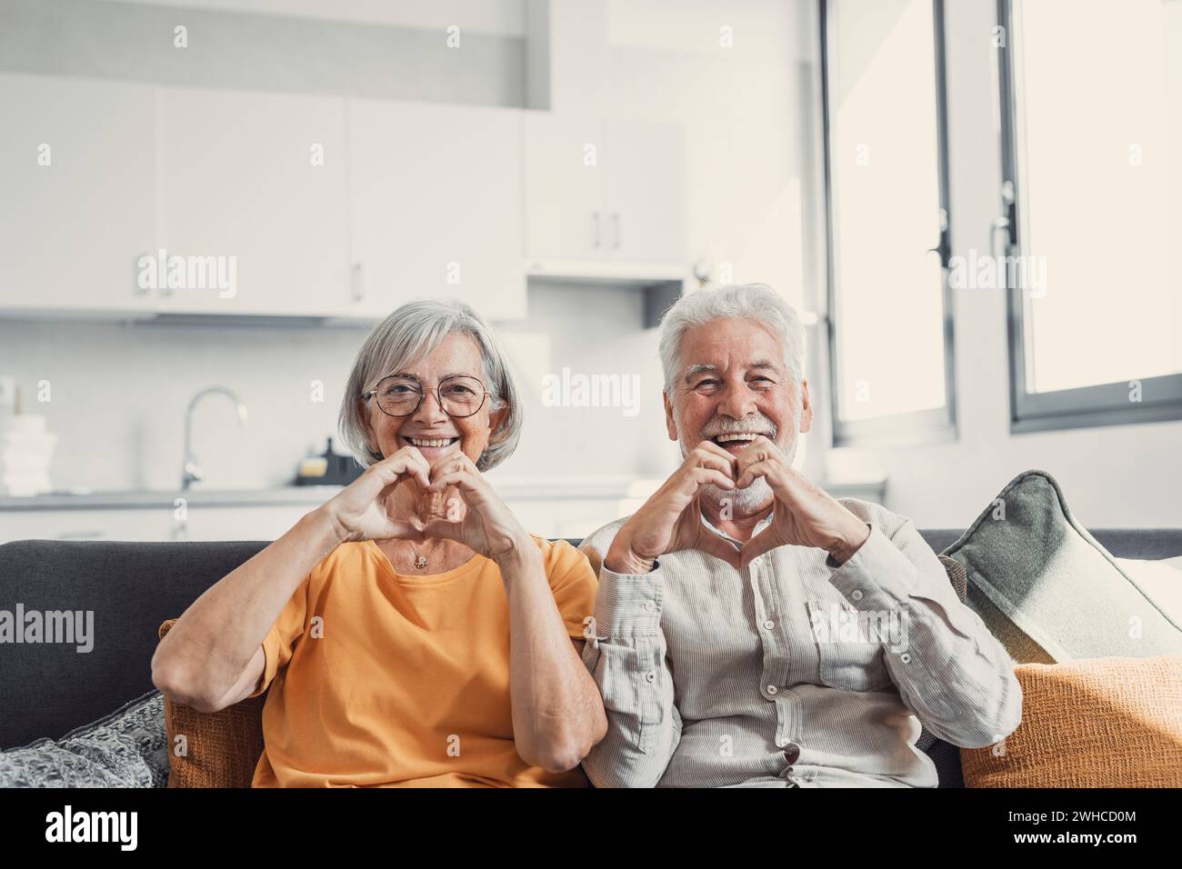 Close up portrait happy sincere middle aged elderly retired family couple making heart gesture with fingers, showing love or demonstrating sincere feelings together indoors, looking at camera. Stock Photo