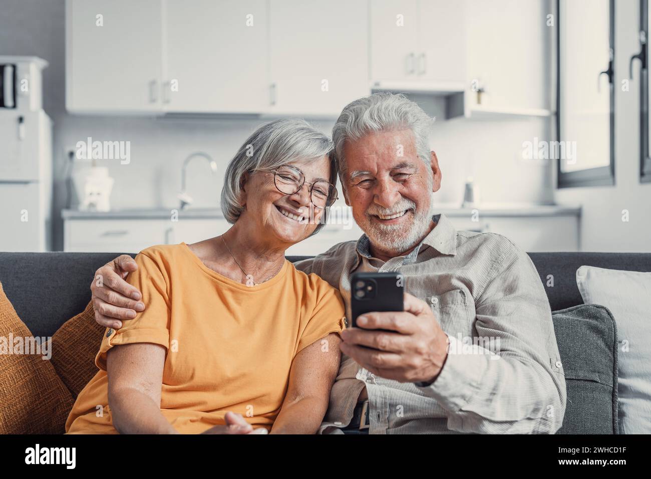 Happy mature family, wife and husband using phone together at home, smiling middle aged man and woman sitting on couch, using mobile device apps, watching video in social network, surfing internet Stock Photo