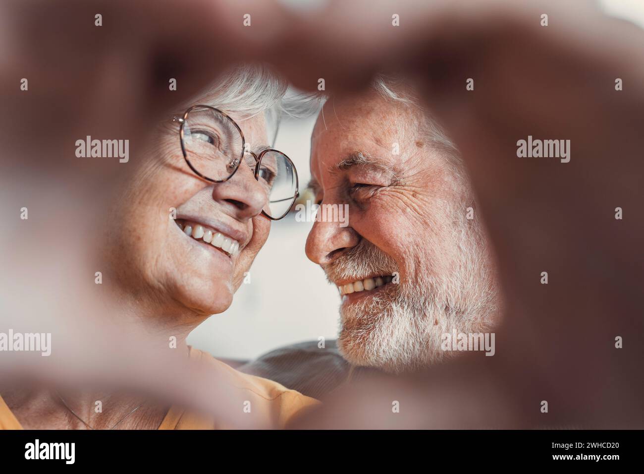 Close up portrait happy sincere middle aged elderly retired family couple making heart gesture with fingers, showing love or demonstrating sincere feelings together indoors, looking at camera. Stock Photo