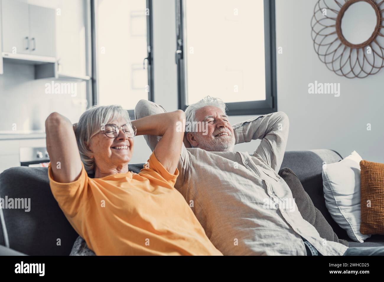 Peaceful middle aged man and woman with closed eyes relaxing on comfortable couch at home, mature family daydreaming together, grey haired wife and husband resting with hands behind head, breathing Stock Photo