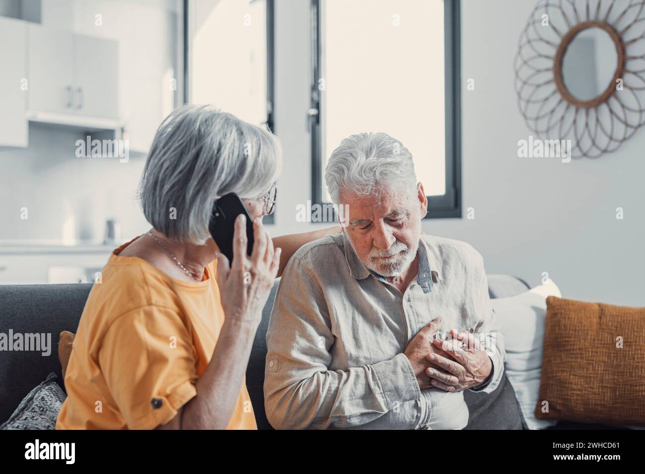 Mature woman, wife calling emergency, talking on phone, holding old husband hand, grey haired man having heart attack, touching chest, suffering from heartache disease at home, feeling pain Stock Photo