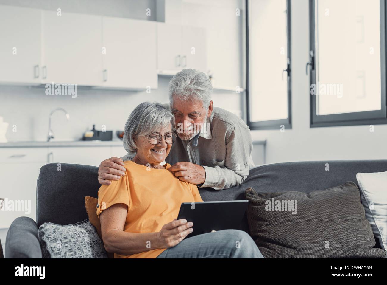 Happy older couple using computer tablet together at home, excited mature man and woman looking at mobile device screen, shopping or chatting online, sitting on cozy couch in living room Stock Photo