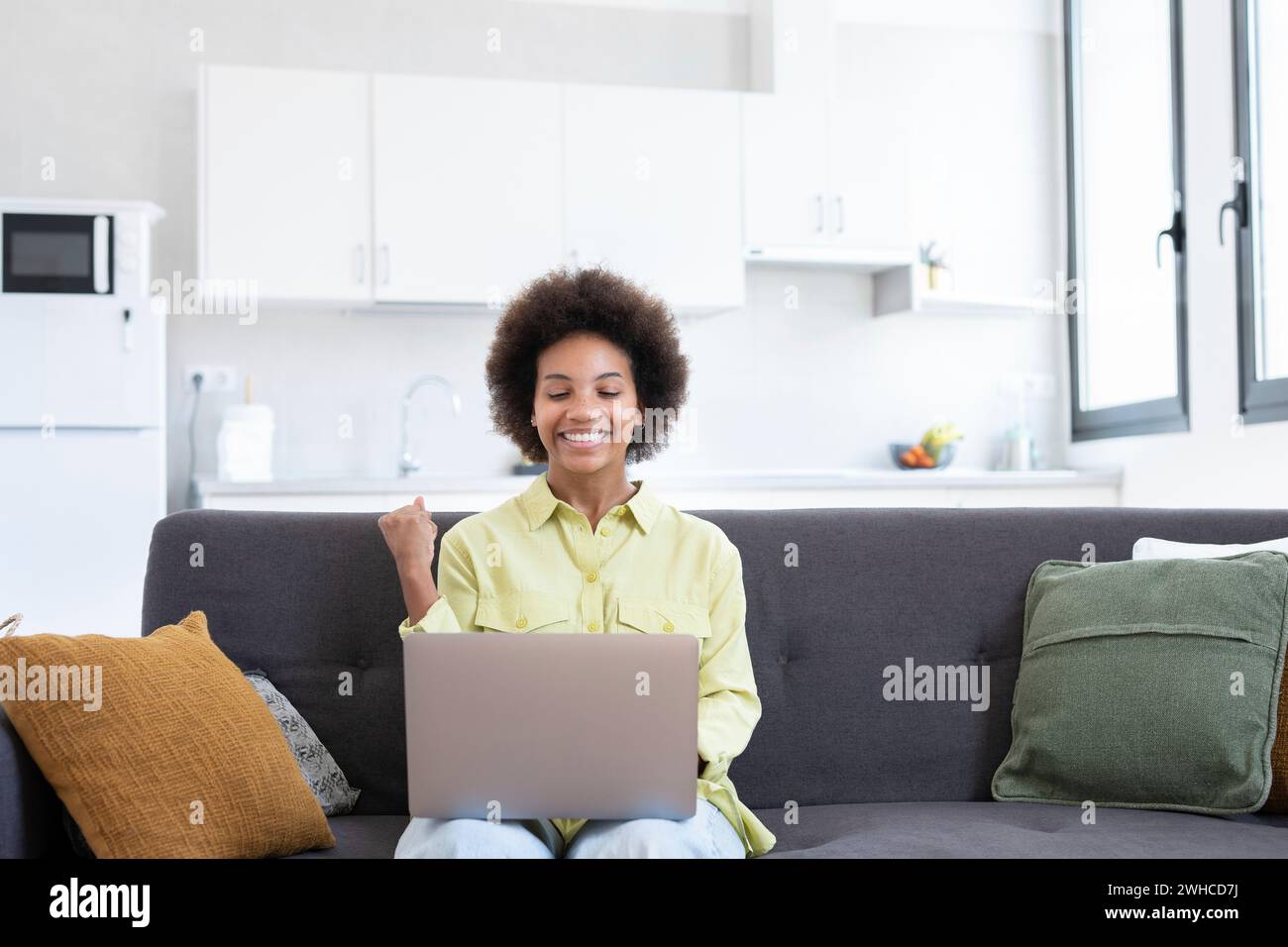 Excited cheerful young Black woman using laptop computer on sofa at home, getting good news, feeling joy, dancing with hands, singing, laughing, making winner gesture, happy to win prize Stock Photo