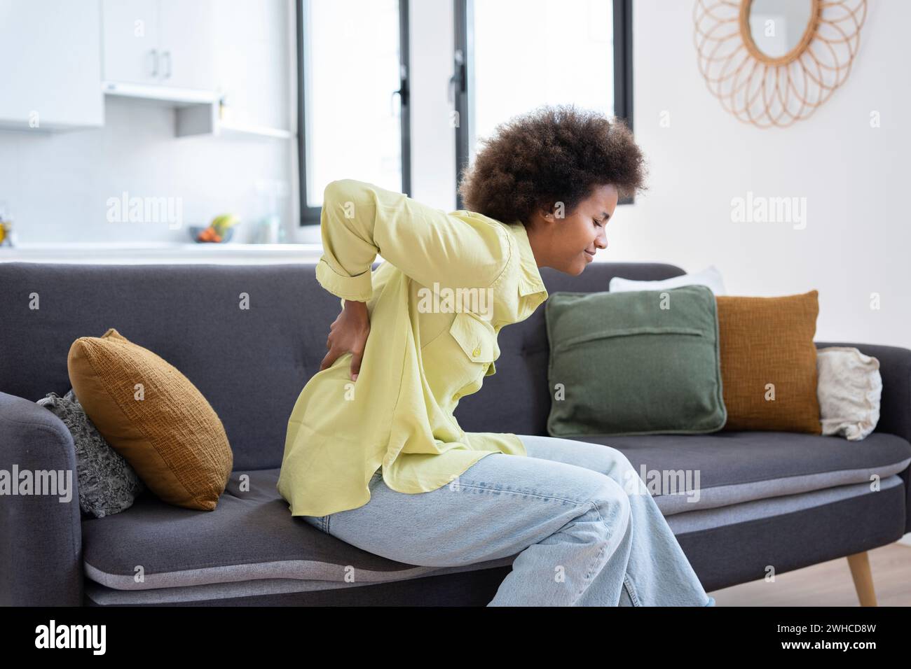 Afro student girl suffers from backache feeling badly, sitting on couch in living room at home. Has a back pain after studying at the computer for a long time. Sedentary work and bad posture concept Stock Photo