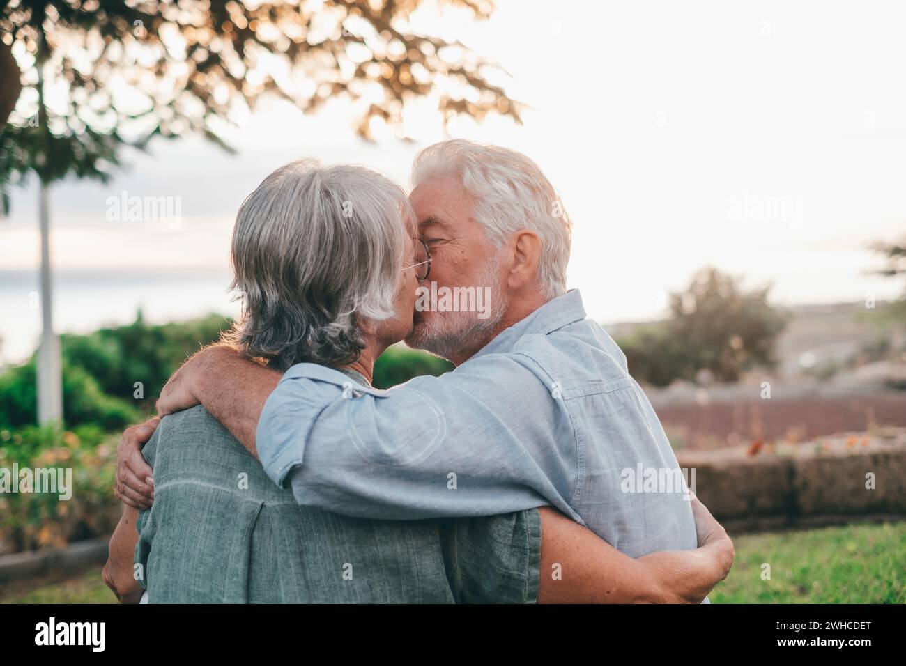 Head shot close up portrait happy grey haired middle aged woman snuggling to smiling older husband, enjoying tender moment at park. Bonding loving old family couple embracing, looking sunset. Stock Photo